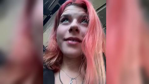 Media: A video of a young woman with pink hair, fair skin, and a silver necklace, smiling and looking upwards, set against a blurred background.