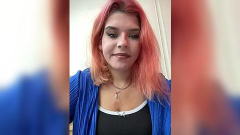 Media: Video of a young woman with light skin and shoulder-length, vibrant pink hair, wearing a blue cardigan over a black top, looking down.