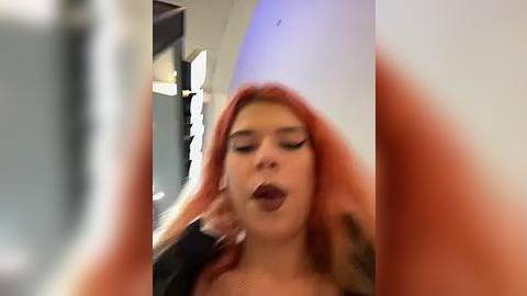 Media: A video of a young woman with long, dyed orange hair, wearing a black top, and a large, white cylindrical hat. She is seen in an indoor setting, possibly a party or event, with blurred background.