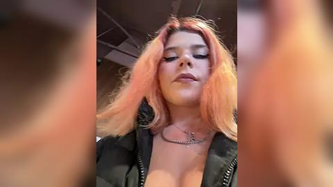 Media: Video of a young woman with shoulder-length, pink-dyed hair, wearing a black leather jacket, and a silver chain necklace. She has light skin and is indoors, with a blurred background.