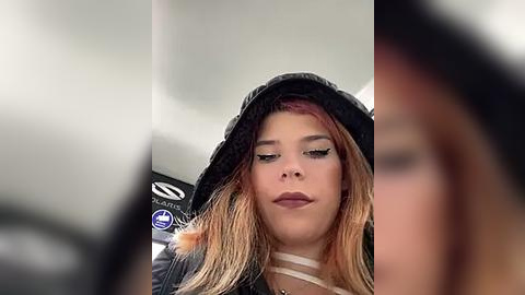 Media: Video of a young woman with long, wavy, light brown hair wearing a black knitted hat and a black jacket. She has light skin and wears makeup with dark eyeliner. The background is blurred, possibly a car interior.