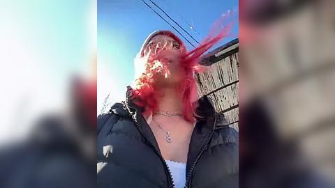 Media: Video of a person with bright red hair, wearing a black jacket and white shirt, captured from a low angle, standing in front of a wooden fence on a clear, sunny day.