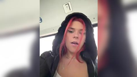 Media: Video of a young woman with fair skin and bright red hair, wearing a black hoodie and a necklace, seated inside a dimly lit car.