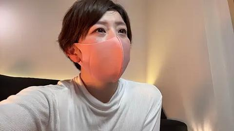 Media: Video of an Asian woman with short dark hair, wearing a white T-shirt and pink surgical mask, looking pensive. Background is blurred with warm lighting.