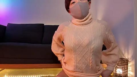 Media: A video of a young Asian woman with short dark hair, wearing a cream-colored knitted sweater, face mask, and a hand on her hip, standing in front of a dark couch and a lit lantern.