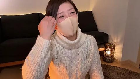 Media: Video of a young Asian woman with short black hair, wearing a beige face mask and a cream-colored knitted sweater, sitting on a beige rug in a dimly-lit living room with a black sofa and a lit candle beside her.