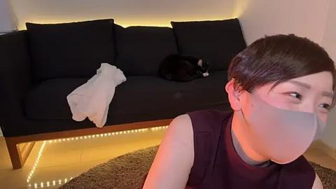 Media: Video of a short-haired Asian woman in a black mask, sitting on a beige rug in a minimalist living room. A black cat sleeps on a dark sofa, with a white towel nearby.