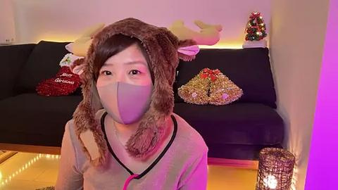 Media: Video of an Asian woman in a fuzzy brown reindeer mask and mask, wearing a pink face covering, sitting on a black couch with festive decorations, including a red Christmas pillow and a gold-wrapped present.