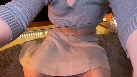 Media: Video of a woman wearing a grey ribbed sweater and a beige skirt, kneeling on a shaggy carpet in a dimly lit room with wooden floors and a glass table.