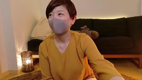 Media: Video of a young Asian woman with short brown hair wearing a mustard-yellow shirt, gray face mask, and a necklace, sitting on a beige rug in a cozy living room with a dark couch and a lit candle in a lantern.