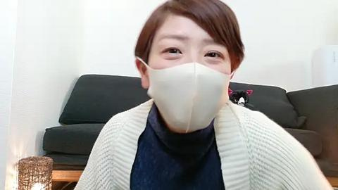 Media: Video of an Asian woman with short brown hair, wearing a beige face mask, navy sweater, and a light-colored cardigan, seated in a living room with a dark couch and wicker basket.