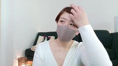 Media: A video of an Asian woman in a white cardigan, wearing a face mask, touching her forehead in a living room with a dark sofa, a blanket, and a lit candle.