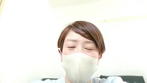 Media: Video of an Asian woman with short brown hair wearing a white surgical mask, eyes closed, in a bright, sterile room with white walls and a black couch in the background.