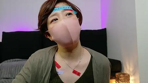 Media: Video of a woman with short brown hair, wearing a mask with a blue headband, green cardigan, and red and white tape over her mouth, in a modern, dimly lit bedroom with a black bed and purple accent lighting.