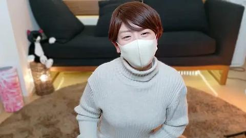 Media: A video of an Asian woman with short brown hair, wearing a beige turtleneck sweater, white face mask, and white gloves, standing in a modern living room with a black sofa, decorative cat figurine, and a brown rug.