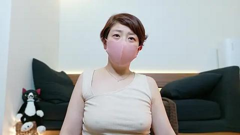 Media: Video of a light-skinned Asian woman with short brown hair, wearing a beige tank top, pink mask, and seated indoors. Background includes a black sofa, plush cat toy, and white walls.