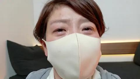 Media: Video of an Asian woman with short brown hair, wearing a beige face mask, looking concerned, in a modern bedroom with black and white decor.