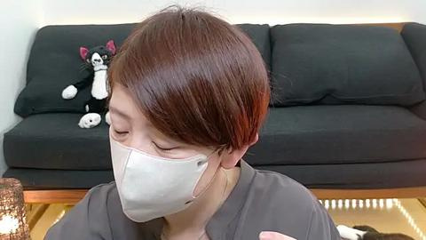 Media: Video of a woman with short, straight brown hair, wearing a white face mask, gray shirt, and sitting on a couch with a black cushion. Background includes a plush black cat toy and a wooden floor.