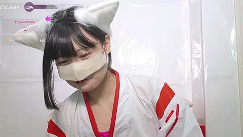 Media: Video of a young East Asian woman in a white hospital gown with red accents, wearing white cat ears, a surgical mask, and a pink shirt, standing in a sterile, white room with a \"Giraffe\" rating display.