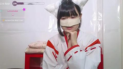 Media: A video of a Japanese woman in a white kimono with red accents, wearing cat ears and a surgical mask, sitting on a red stool in a sterile room.