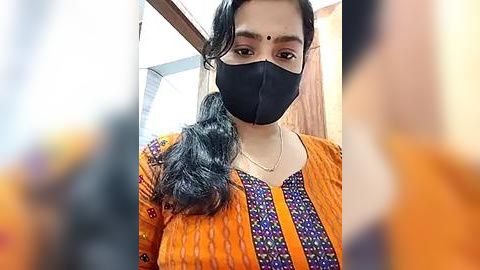 Media: Video of a South Asian woman with a fair complexion, wearing a black face mask, orange saree with floral patterns, and a gold necklace. She has long, wavy black hair and a bindi on her forehead. Background shows blurred, out-of-focus people.