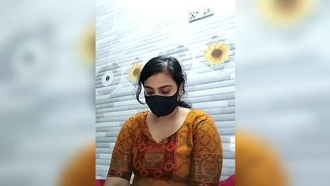 Media: Video of a woman in an orange floral blouse, black mask, and long dark hair, sitting against a white brick wall adorned with sunflower decals, indoors.