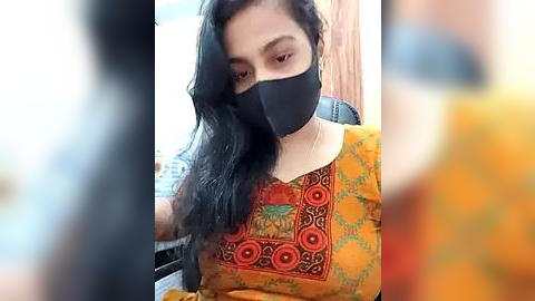 Media: Video of a South Asian woman with long black hair, wearing a black face mask, orange sari with intricate embroidery, sitting indoors. Background blurred.