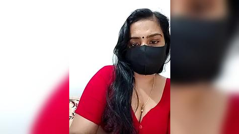 Media: Video of a South Asian woman with long black hair, wearing a red blouse, black face mask, and beige necklace, set against a blurred background.