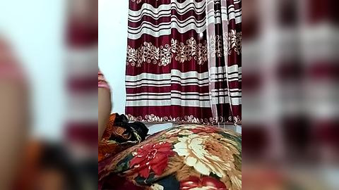 Media: Video of a woman with medium skin tone and dark hair, wearing a maroon and white floral-patterned sari, seated on a bed with a colorful, intricate floral-patterned quilt. The background is blurred, focusing on the subject.