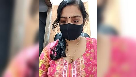 Media: Video of a woman with medium brown skin, wearing a black face mask, pink and yellow traditional top, and a gold necklace, seated indoors, blurred background.