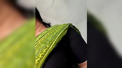 Media: Video of a woman wearing a green, embroidered sari over a black top, partially covered by a white shawl, blurred background.