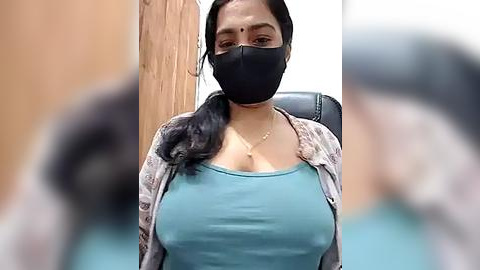 Media: A video of a South Asian woman with medium brown skin, wearing a black mask, blue tank top, and patterned cardigan, seated indoors, with blurred background.