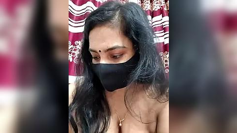 Media: Video of a South Asian woman with long black hair, wearing a black face mask, a red bindi, and a gold necklace, standing in front of a maroon curtain with white floral patterns.