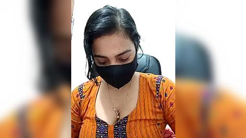 Media: Video of a woman with black hair wearing a black face mask, orange and purple traditional attire, seated indoors.