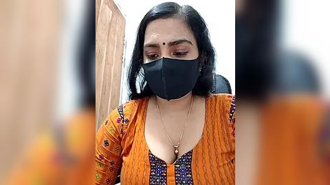 Media: Video of a woman with dark hair, wearing an orange floral blouse, black mask, and a black bindi, sitting indoors against a blurred wooden backdrop.