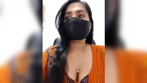 Media: Video of a young woman with long black hair, wearing a black face mask and orange top, sitting against a plain white background.