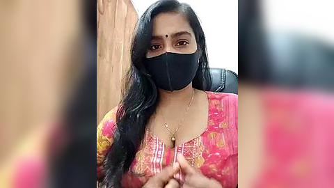 Media: Video of a South Asian woman with long black hair, wearing a pink floral blouse, black face mask, and a gold necklace, seated in a wooden room.