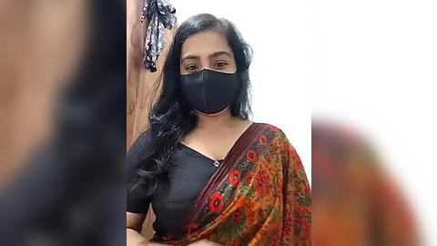 Media: Video of a woman with long black hair, wearing a black mask, black top, and colorful sari, standing indoors.