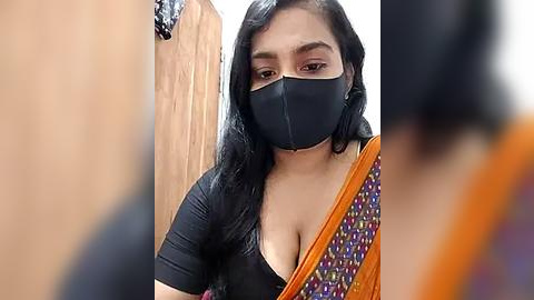 Media: Video of a South Asian woman with long black hair, wearing a black mask, black top, and a colorful sari, standing indoors near a wooden door.