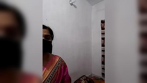 Media: A video showing a woman in a maroon sari with gold trim, partially covered by a black mask, standing in a small, plain white room with a patterned curtain and a mirror reflecting her.