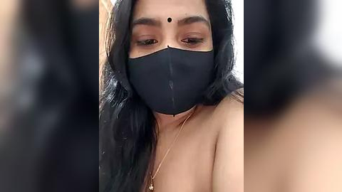 Media: A close-up video of a South Asian woman with long black hair, wearing a black mask, gold necklace, and a traditional bindi, with a blurred background.