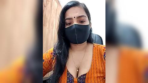 Media: Video of a South Asian woman with long black hair, wearing a black mask, orange blouse, and a bindi, seated indoors with blurred background.