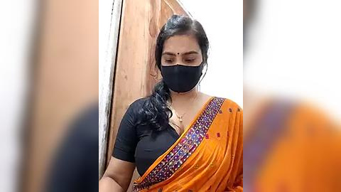 Media: Video of a South Asian woman in a black mask, orange saree with floral embroidery, and black top, standing in a wooden doorway. Background features blurred figures.