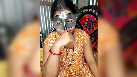 Media: Video of a young woman with dark skin, wearing an orange-patterned blouse, silver masquerade mask, and red bangles, seated on a colorful cushioned chair.