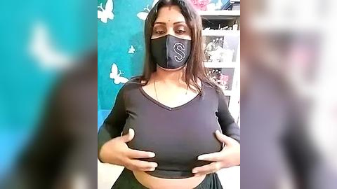 Media: A video of a young woman with medium skin tone, wearing a black face mask and black long-sleeve top, holding her breasts, against a teal wall with butterfly decals and bookshelves.