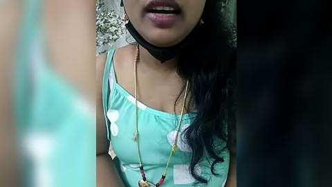 Media: Video of a woman with medium brown skin, wearing a turquoise tank top with white polka dots, a black choker, and a long necklace with a red pendant. She has long, dark hair and is smiling.