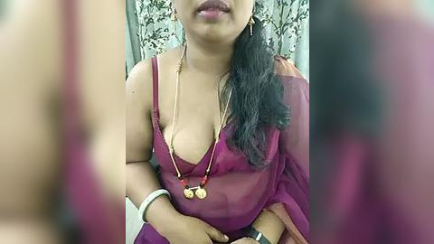 Media: Video of a South Asian woman with medium skin tone, wearing a sheer maroon sari, showing cleavage, gold necklace, and wristwatch. Background features floral-patterned curtains.