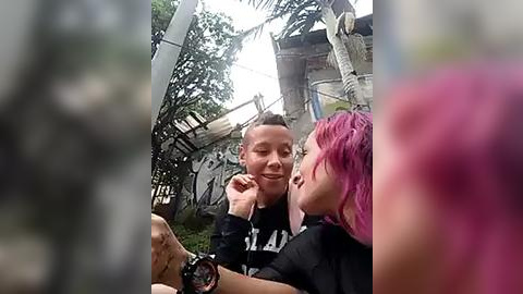 Media: A video captures two people with pink hair smiling at each other in a jungle-themed amusement park. The man, with short hair, wears a black t-shirt; the woman, with long hair, wears a black top. The background features wooden structures and greenery.