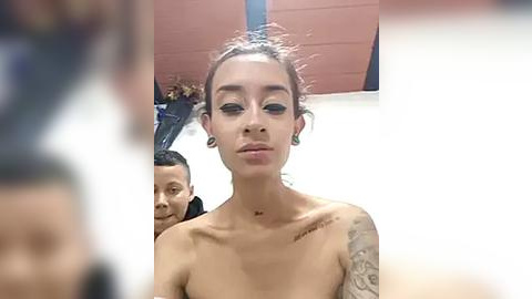 Media: Video of a topless, light-skinned woman with medium-length, tied-back hair, tattoos on her shoulder and neck, standing indoors.