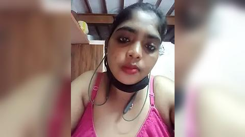 Media: Video of a young South Asian woman with medium brown skin, wearing a black choker, pink tank top, and headphones, indoors with wooden furniture.